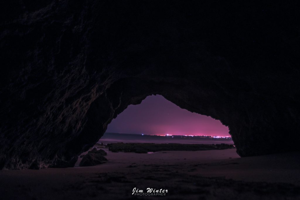 Arche rocheuse en bord de mer de nuit ouvrant sur un ciel rose et des lumières lointaines