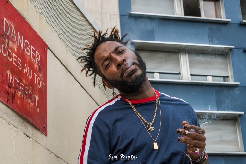 Homme à dreadlocks fumant contre un mur portant un panneau rouge Danger, portrait urbain